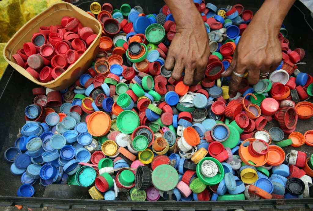 A pair of hands sorts through a collection of bottle caps|Plants grown in upcycled plastic bags|An old plastic barrel upcycled into a water dispenser|Plastic poms poms upcycled into a table decoration|A plastic barrel upcycled into a plant pot with a bush growing out of it|Upcycled plastic waste that has been turned into a butterfly shaped decoration|A plastic barrel upcycled into a plant pot|A plastic barrel upcycled into a water dispenser|An illustration of the basic idea behind Google Task Mate