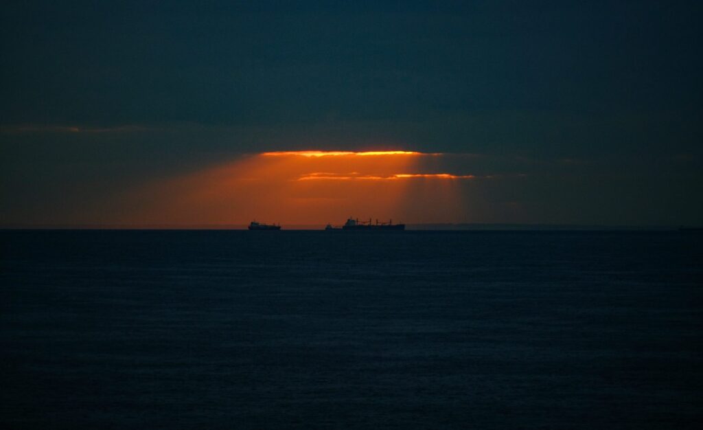 Freighter ships off the coast of the UK are in the distance silhouetted against bright orange light breaking in from the clouds.