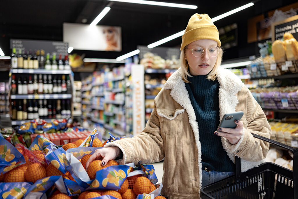 Young woman using cell phone while buying fresh oranges in the supermarket.
